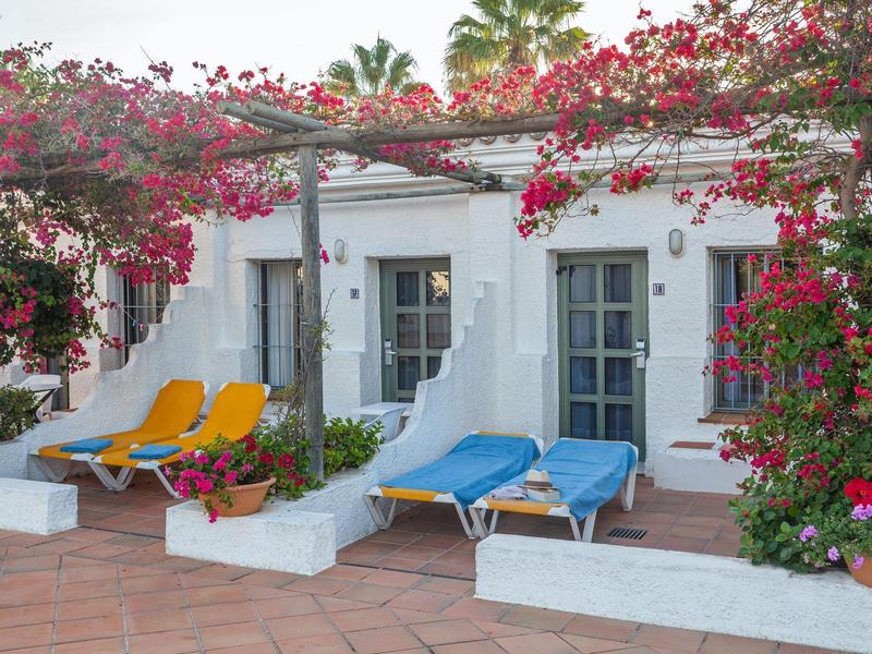 Terrazza con lettini colorati sotto pergolato fiorito di fronte a camere bianche.