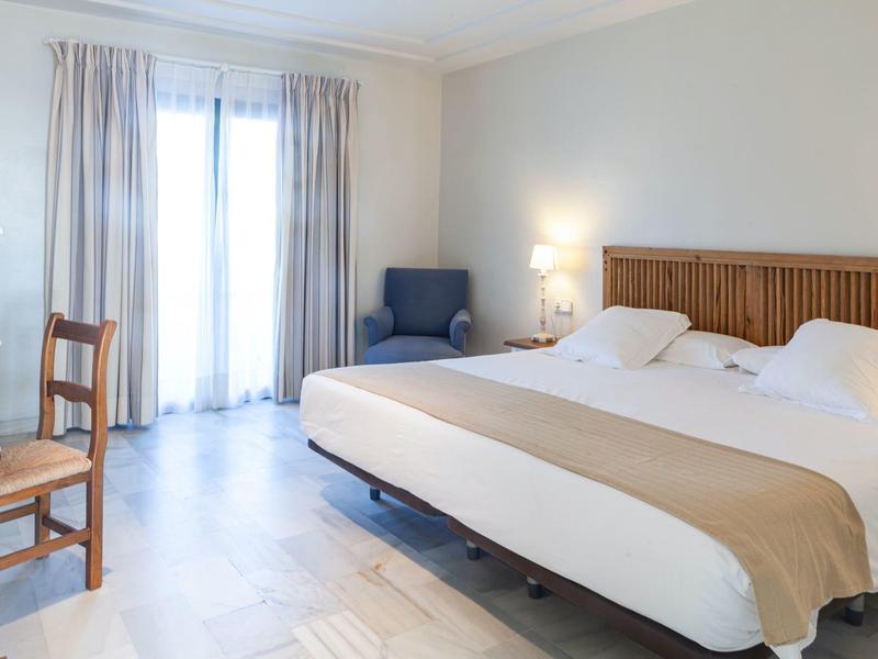 Camera d'albergo luminosa con letto grande, poltrona blu e scrivania in legno vicino alle finestre.