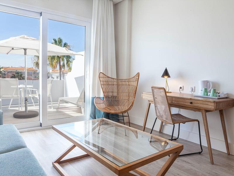 Soggiorno luminoso con poltrona in vimini, scrivania in legno e terrazza con ombrellone.