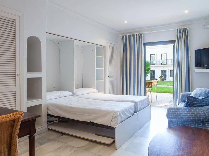 Camera d'hotel luminosa con letto matrimoniale, poltrona blu, scrivania e accesso al giardino.