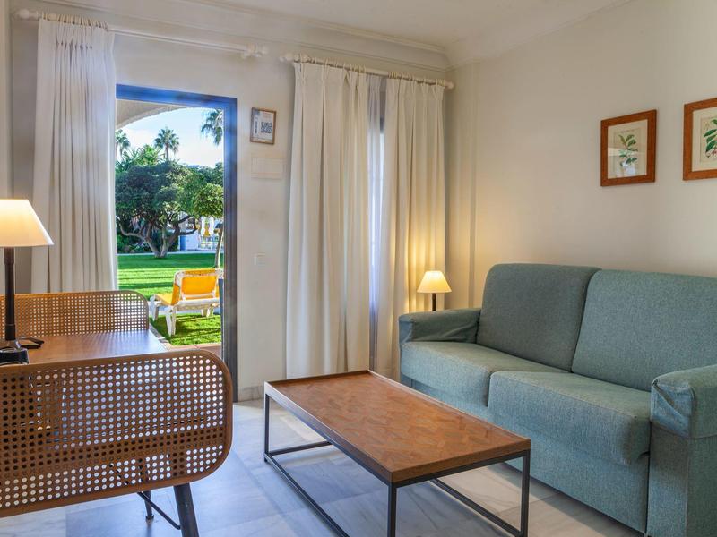 Soggiorno luminoso con divano verde, tavolino e scrivania in legno davanti a una porta che si apre su un giardino.