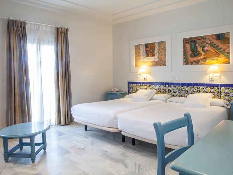 Camera d'hotel luminosa con due letti singoli, mobili blu e due quadri alle pareti.