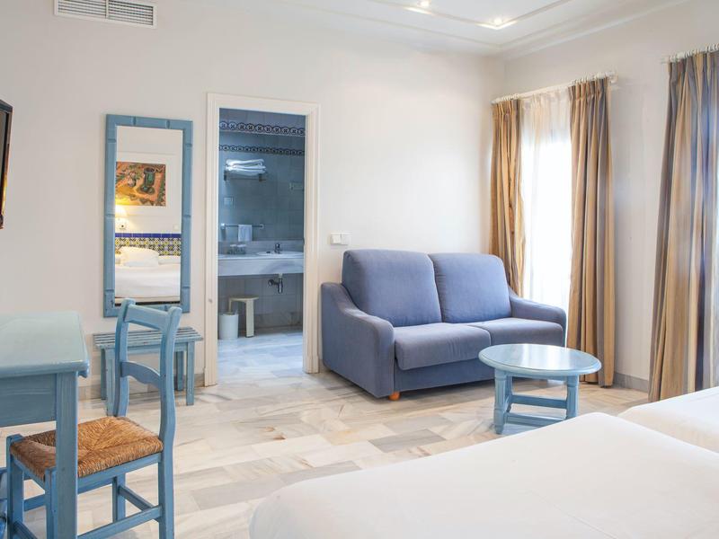 Camera d’albergo luminosa con divano, tavolo, sedie blu e grandi finestre.