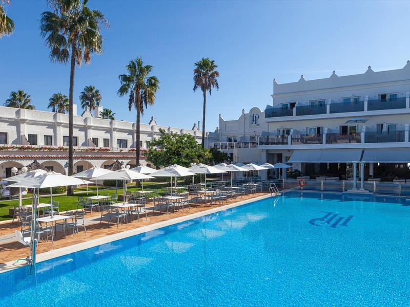 Piscina azzurra chiara circondata da lettini e ombrelloni vicino a un hotel bianco.