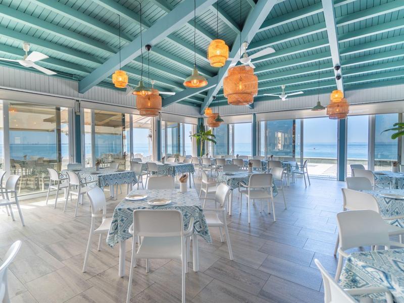 Ristorante luminoso con vista mare, tavoli e sedie moderne, atmosfera ariosa e decorazioni arancioni.