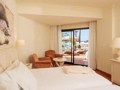 Hell eingerichtetes Hotelzimmer mit Doppelbett, Sesseln und Zugang zu einer Terrasse mit Blick auf den Pool.