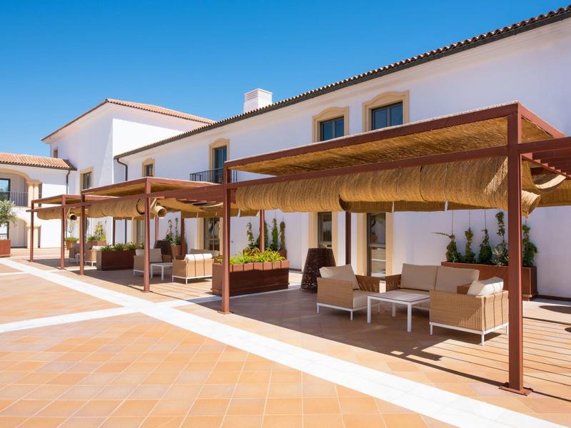 Lichte buitenlounge met houten pergola en comfortabele zitplaatsen voor een wit hotelgebouw.