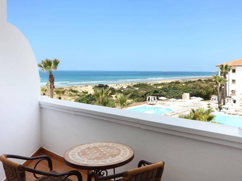 Balkon met tafel en twee stoelen met uitzicht op zwembad, palmbomen en zee onder een heldere hemel.