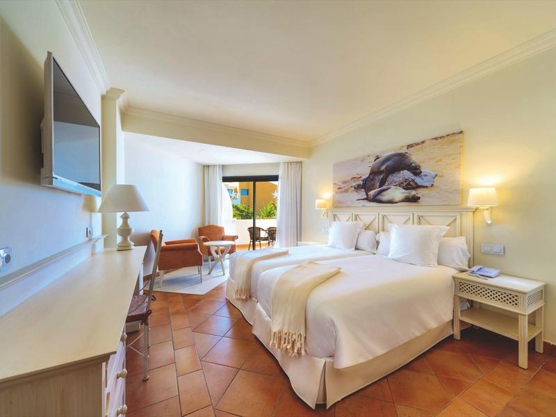 Lichte hotelkamer met tweepersoonsbed, tv, stoel en balkon met uitzicht op groen.