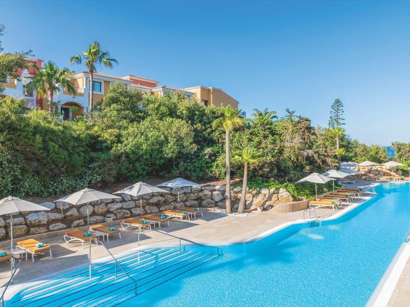 Een groot buitenzwembad met ligstoelen en parasols naast groene begroeiing en een hotelgebouw op de achtergrond.