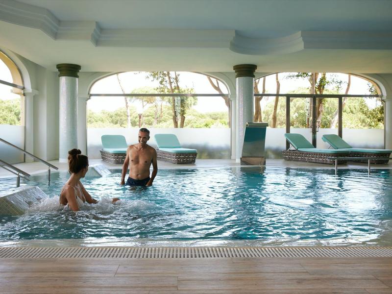 Binnenzwembad met zuilen en grote ramen, twee mensen ontspannen in het water.