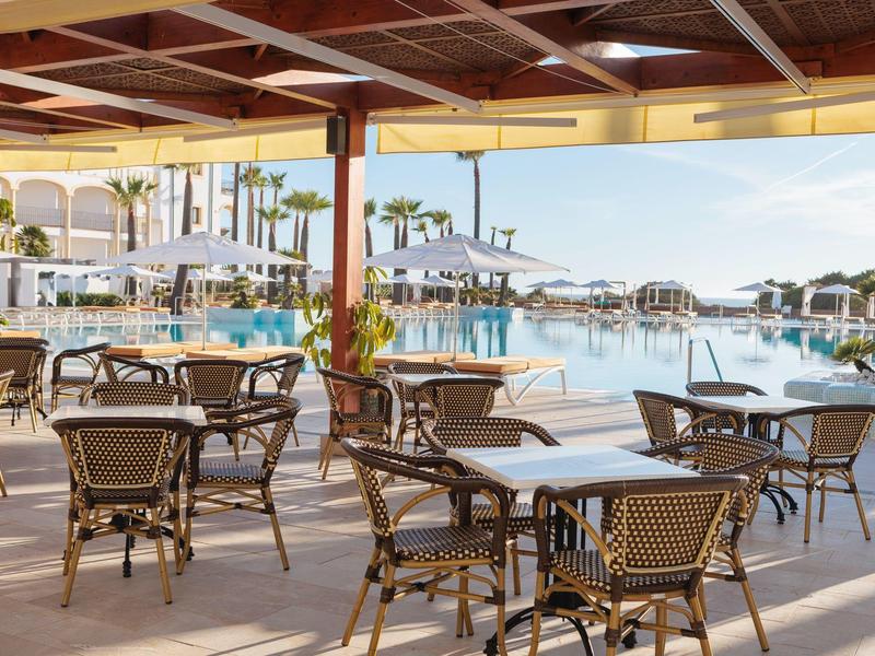 Overdekt terras met tafels en stoelen naast een hotelzwembad en parasols bij helder weer.