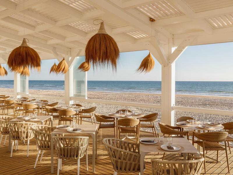 Open strandrestaurant met ronde tafels, touwstoelen en strooien lampen met uitzicht op de zee.