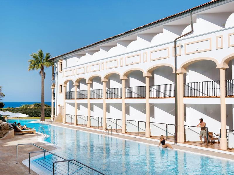 Hotel met groot buitenzwembad, palmbomen en uitzicht op zee op een zonnige dag