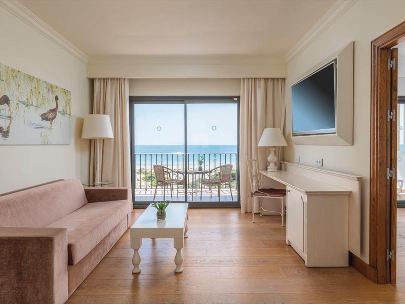 Lichte hotelkamer met zeezicht, bank, tafels en balkon met stoelen.