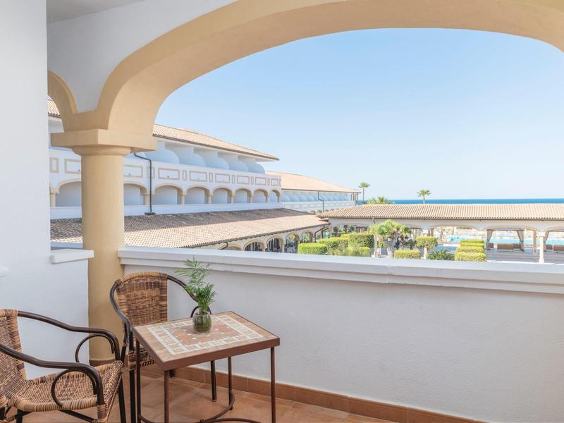 Balkon met tafel, stoelen en uitzicht op hotelterrein en zee onder blauwe lucht.