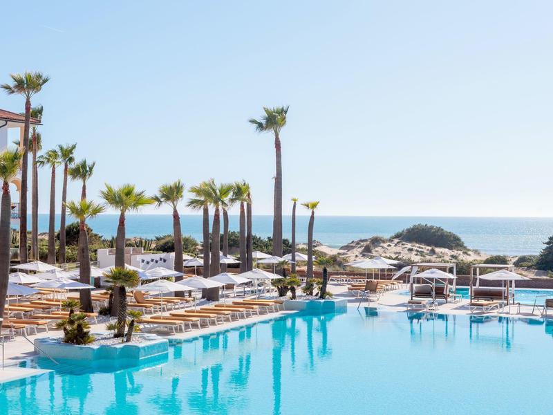 Een hotelzwembad met palmbomen voor de zee onder een heldere hemel.