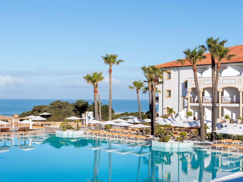 Hotel met groot zwembad, ligstoelen en palmbomen met uitzicht op zee onder een heldere hemel.