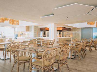 Helles, modernes Restaurant mit Holztischen, geflochtenen Stühlen und Deckenlampen in warmem Licht.