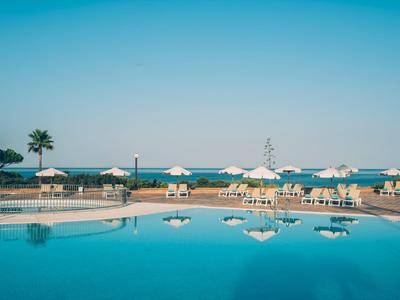 Großer Pool mit weißen Sonnenschirmen und Palmen, darunter klarer blauer Himmel und Meer im Hintergrund.