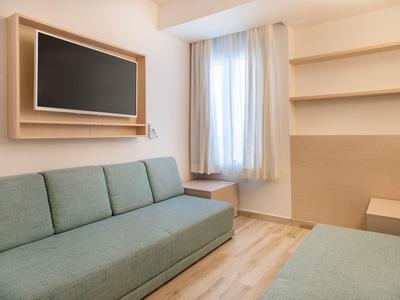 Kleines Wohnzimmer mit grünem Sofa, Holzregalen, Fenster mit Vorhang und hellem Holzboden.