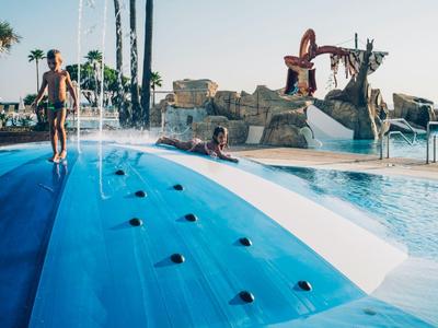 Kinder spielen auf einer blauen Wasserrutsche mit Wasserspritzern im Schwimmbad bei Sonnenschein.