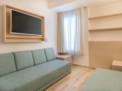 Kleiner heller Wohnraum mit grünem Sofa, Wand-TV und zwei Holztischen neben Fenster mit weißen Gardinen.
