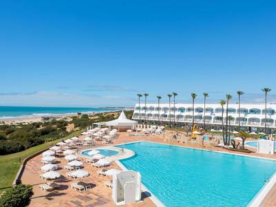 Hotelpool mit blauer Wasserfläche, weißen Liegen und Palmen vor dem blauen Himmel am Meer.