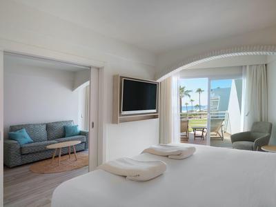 Licht hotelkamer met bed, bank, tv en balkon met uitzicht op palmbomen.