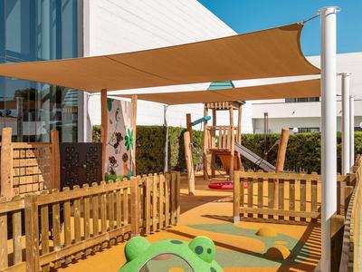 Moderner Außen-Spielplatz mit Kletterwand, Holzzaun und Sonnensegel neben einem Hotelgebäude.
