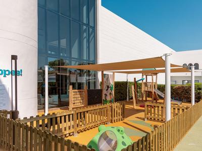 Moderner Hotelspielplatz mit Holzzäunen und Sonnenschutz unter blauem Himmel.