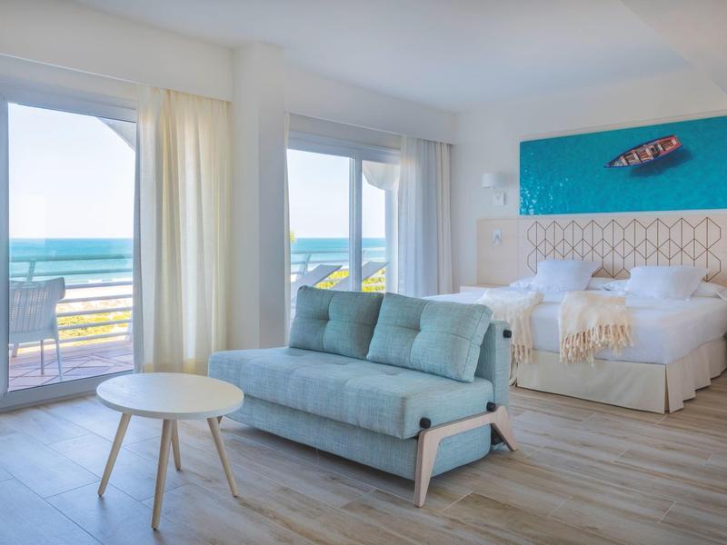 Lichte hotelkamer met tweepersoonsbed, bank en zeezicht door grote ramen.