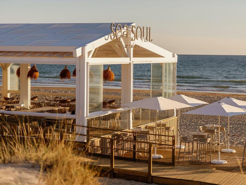 Witte paviljoen aan het strand met zitplaatsen en parasols bij rustige zee tijdens zonsondergang.
