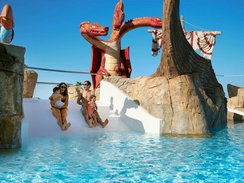 Kinderen glijden van een glijbaan in een zwembad onder een heldere blauwe lucht.