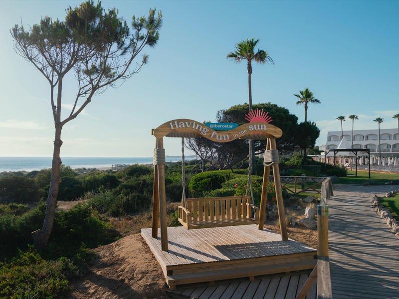 Houten schommel op speeltuin bij strandpad, omgeven door bomen, met uitzicht op zee.