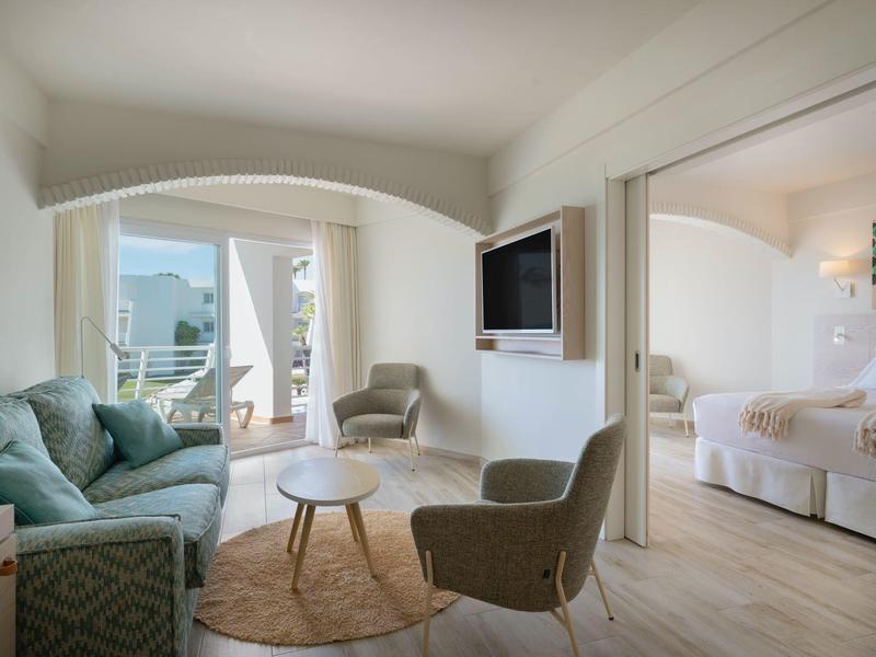 Lichte woonkamer met bank, fauteuils, flatscreen-tv, aangrenzende slaapkamer met tweepersoonsbed.