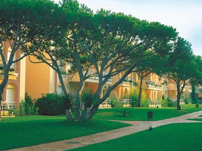 Duże drzewa przed budynkiem hotelu z zadbaną trawą i ścieżkami.