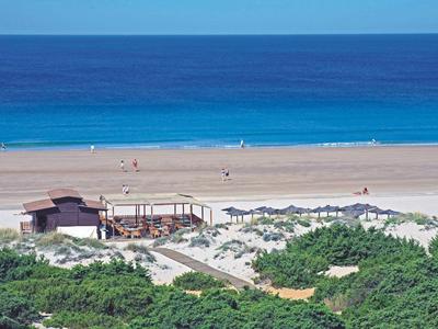 Blauer Ozean, breiter Sandstrand mit Sonnenschirmen, davor grüne Büsche und Holzgebäude.