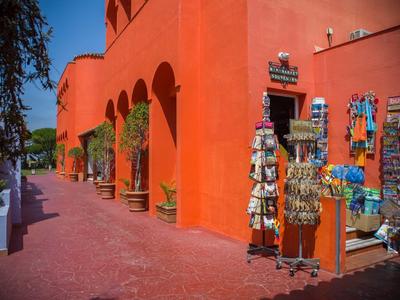 Rotes Gebäude mit Bögen, Souvenirständer und Pflanzen entlang eines gepflasterten Wegs bei sonnigem Wetter.