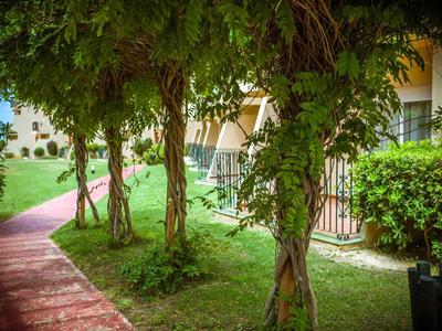 Grüner Gartenweg mit blühenden Bäumen und Sitzbereich neben einem hellen Gebäude.