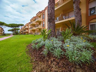 Grüne Pflanzfläche mit Büschen und Bäumen vor gelben Wohngebäuden unter bewölktem Himmel.