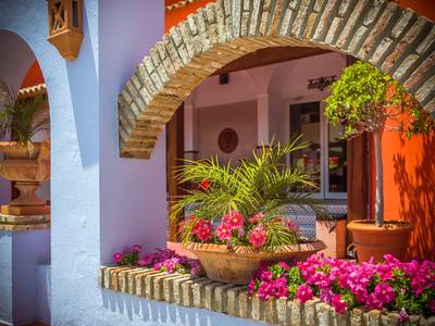 Bunter Innenhof mit Steinbogen, blühenden Blumen, Palmen und Töpfen, helle Wandfarben in Blau und Orange.