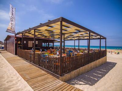 Strandbar aus Holz mit Überdachung, Tischen und Stühlen, umgeben von Sand und blauem Himmel.