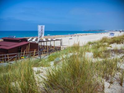 Strand mit Sanddünen, Gras, blauer See, blauer Himmel und roten Holzhütten am Ufer.