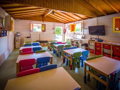 Bunt eingerichteter Klassenraum mit kleinen Tischen, Stühlen, großer Tafel und Fernseher unter Holzdach.