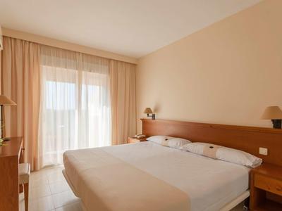 Helles Schlafzimmer mit Doppelbett, Holzmöbeln und großen Fenstern mit Gardinen.