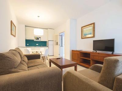 Gemütliches Wohnzimmer mit grauen Sofas, Holztisch, TV und offener Küche im Hintergrund.