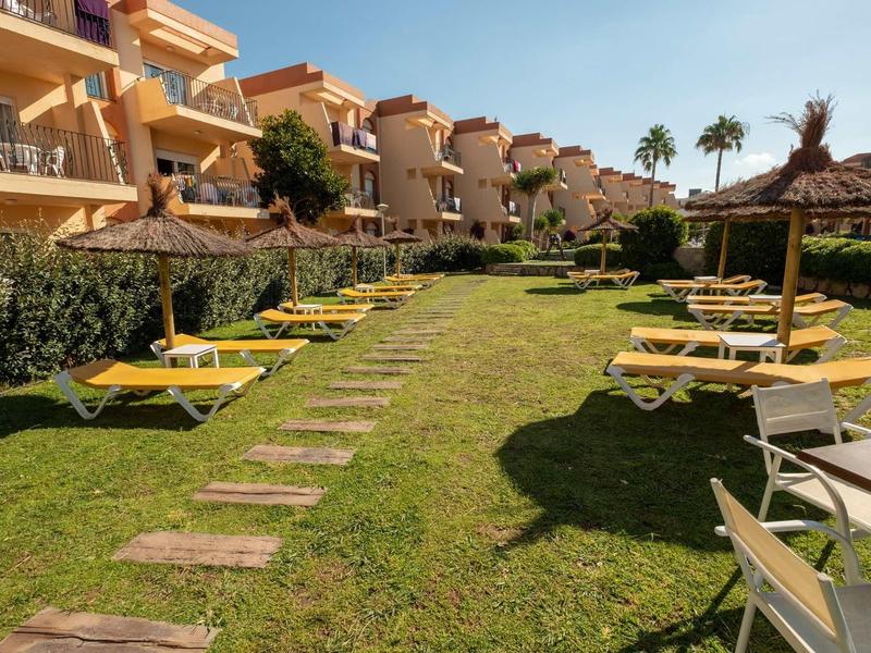 Grüne Liegewiese mit gelben Sonnenliegen und strohgedeckten Sonnenschirmen vor Wohnhäusern.