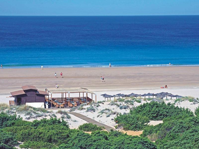 Blauer Ozean, breiter Sandstrand mit Sonnenschirmen, davor grüne Büsche und Holzgebäude.