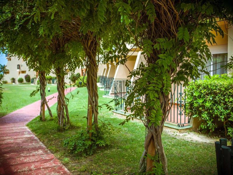 Grüner Gartenweg mit blühenden Bäumen und Sitzbereich neben einem hellen Gebäude.